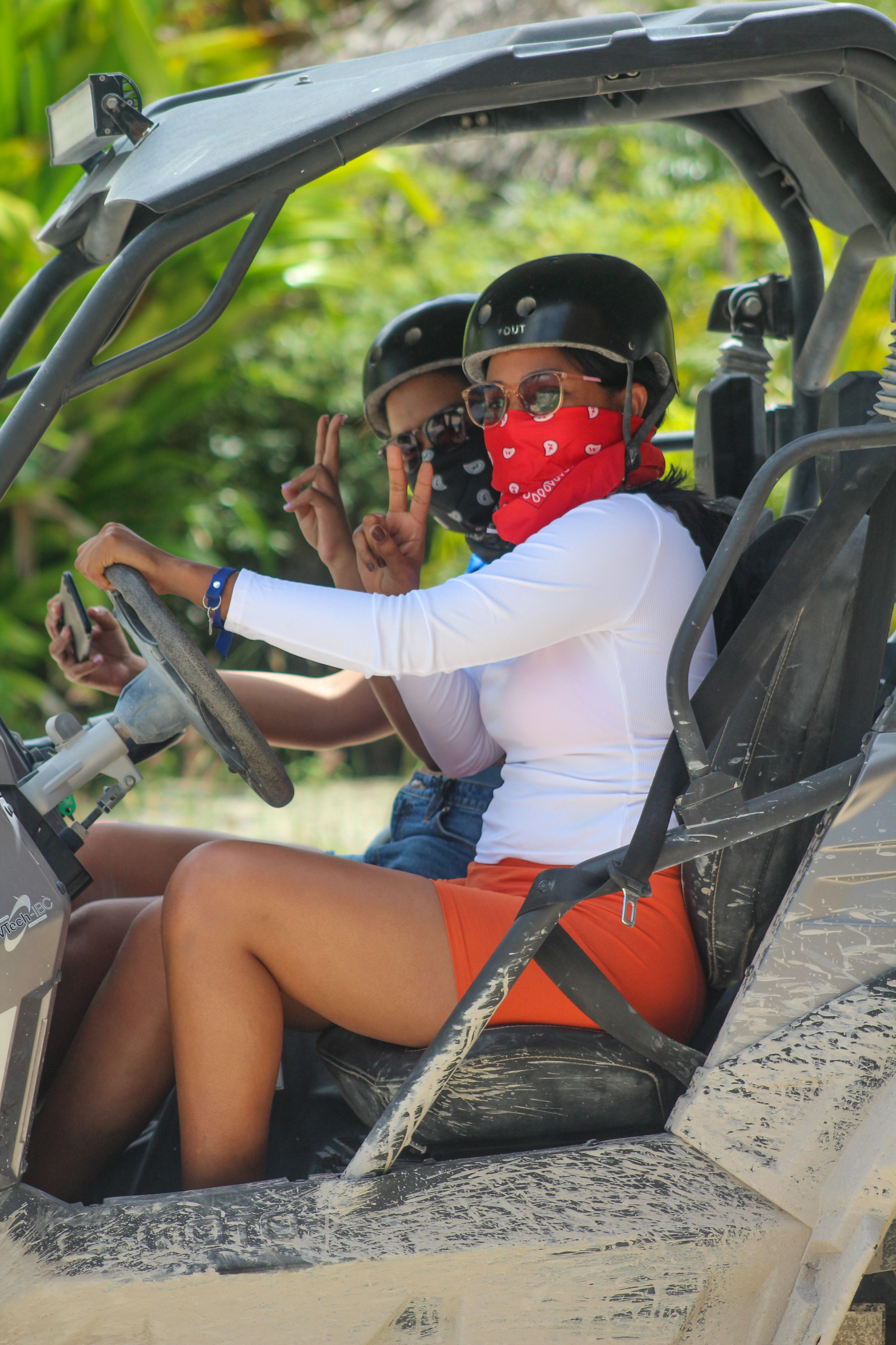Two riders in a muddy off-road UTV amid tropical greenery, wearing helmets, sunglasses and bandana face coverings; driver in white top and orange shorts flashes a peace sign — beach ATV adventure.