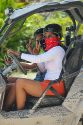 Two riders in a muddy off-road UTV amid tropical greenery, wearing helmets, sunglasses and bandana face coverings; driver in white top and orange shorts flashes a peace sign — beach ATV adventure.