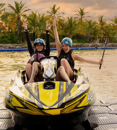 Two smiling riders in blue helmets cheering on a bright yellow jet ski at sunset in a tropical lagoon lined with palm trees