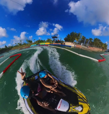 Action shot of a yellow jet ski with two riders in blue helmets speeding across green water at a tropical beach water park, wake trailing past palm trees, inflatable obstacles, and a bright blue sky with puffy clouds.
