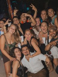 Energetic group of friends at a lively outdoor nighttime party, smiling, posing and holding drinks — fun vacation nightlife scene.