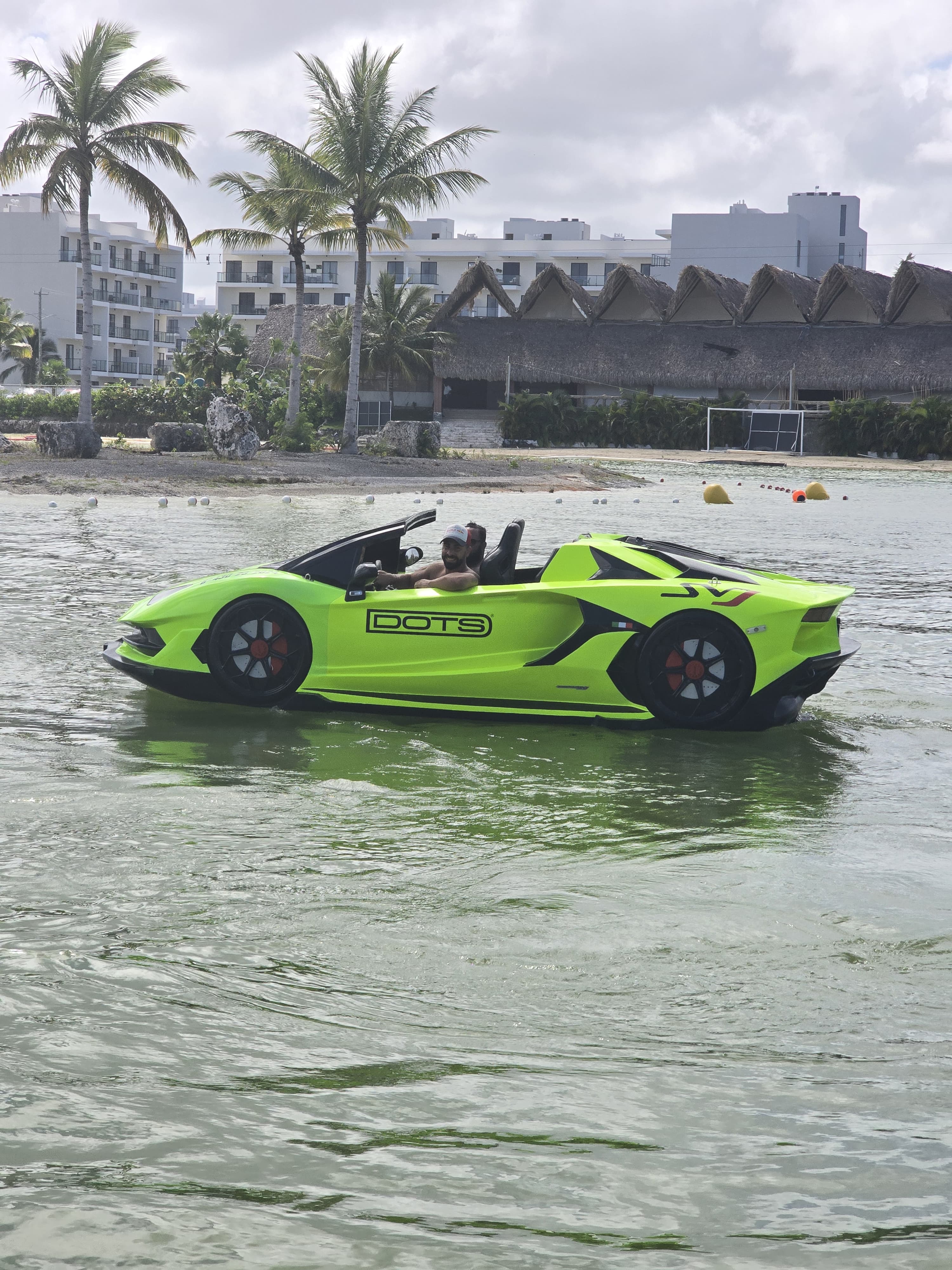Neon-green amphibious sports car gliding in shallow coastal water near a tropical beach resort with palm trees and thatched-roof huts, person at the wheel.