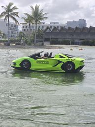 Neon-green amphibious sports car gliding in shallow coastal water near a tropical beach resort with palm trees and thatched-roof huts, person at the wheel.