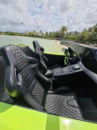 Bright green speedboat interior with black quilted leather seats and neon steering wheel, moored in a shallow tropical lagoon by a palm-lined shoreline under a cloudy sky.