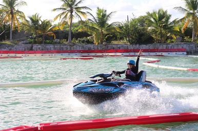 Person on a blue recreational jet ski racing through a tropical lagoon course, splashing turquoise water past red lane barriers with palm trees in the background.