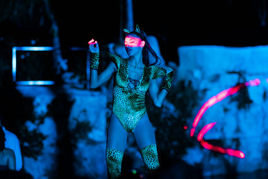 Neon-lit dancer in leopard-print bodysuit and cat ears striking a pose on a dark outdoor stage, bathed in blue light with vivid pink light streaks.