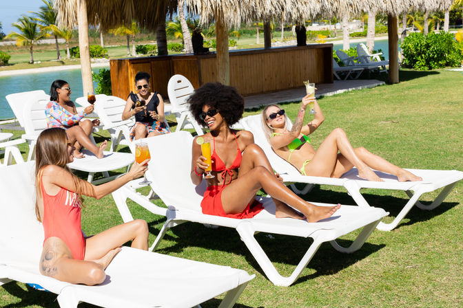 Group of women lounging on white sunbeds under thatched palapas at a tropical resort, toasting colorful cocktails by a lagoon and palm trees.