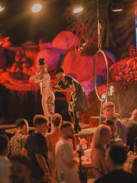 Live body-painting performance on a lit stage at a tropical beach-club style night venue, artist painting a model with black swirl designs beside a suspended aerial hoop while a lively crowd watches under warm red stage lights.