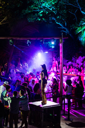 Vibrant tropical outdoor nightclub at night with purple-blue neon lights, a dancer in leopard-print costume posing on a lit platform while a crowded crowd of partygoers dance and drink beneath palm trees.