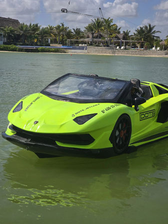 Neon green amphibious convertible sports car floating on a calm tropical lagoon near a palm-lined coastal resort under a sunny sky.