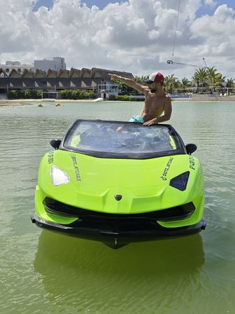 Shirtless man in a baseball cap pointing from a neon green sports-car-style jet boat floating in a tropical resort lagoon with palm trees, thatched roofs and a cloudy sky.