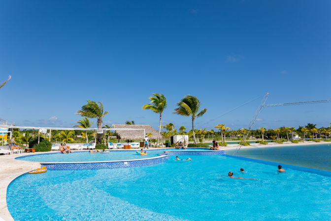 Palm-fringed tropical resort with a large blue infinity-style pool, swimmers and loungers, cabanas and sandy shore under a clear blue sky