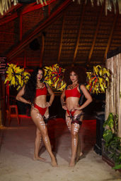 Two carnival dancers in red bikinis with yellow-red feathered wings and white tribal body paint, posing barefoot on sand under a thatched palapa with warm red lighting — tropical beach club, nightlife vibe.