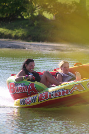 Two friends grinning in life jackets riding a colorful red-and-yellow inflatable towable tube across a sunlit lake with a green shoreline