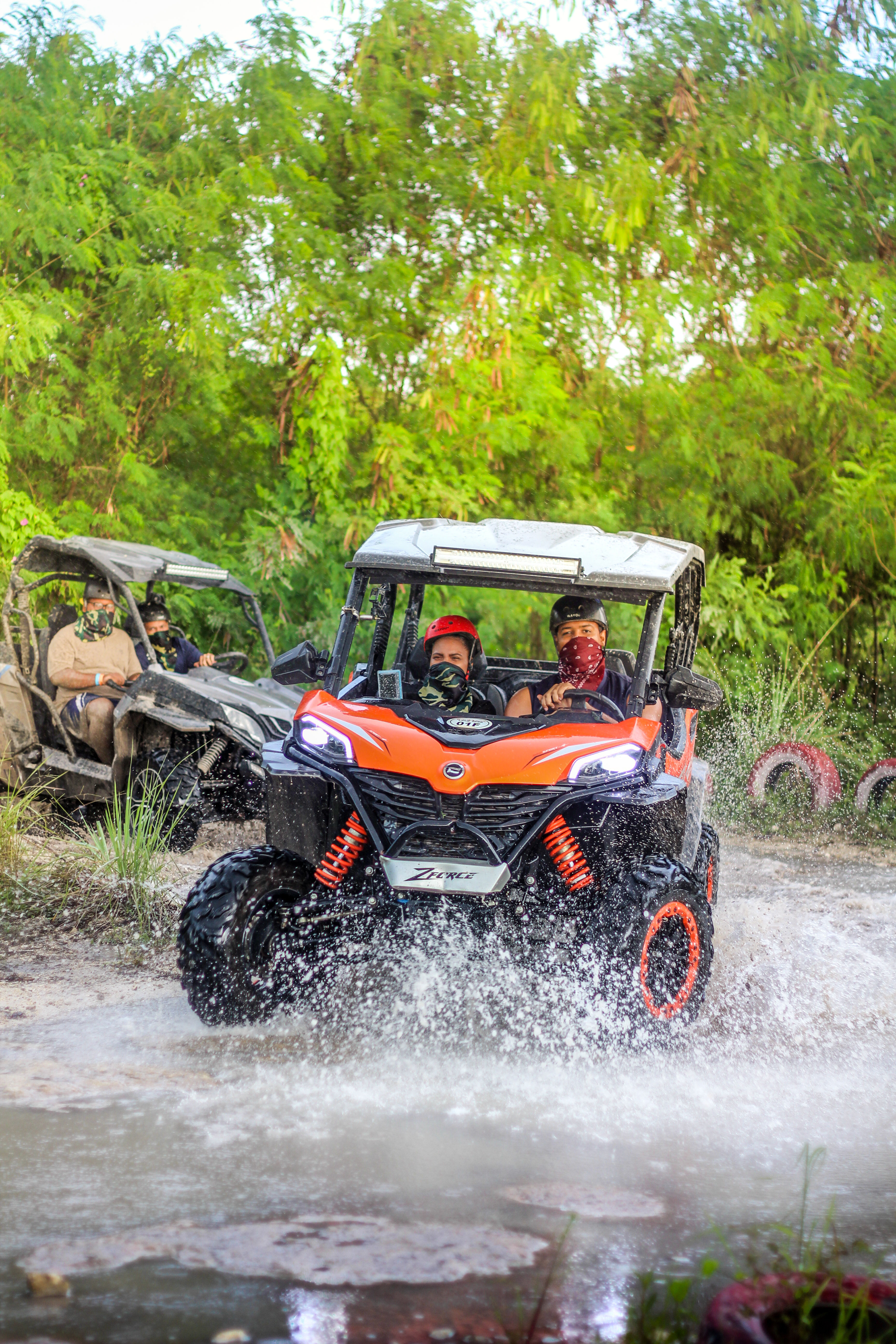 Orange side-by-side UTV splashing through a muddy forest trail with two helmeted riders wearing bandanas and another UTV following amid lush tropical greenery