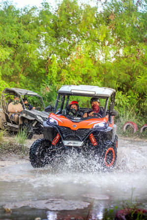 Orange side-by-side UTV splashing through a muddy forest trail with two helmeted riders wearing bandanas and another UTV following amid lush tropical greenery