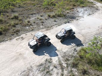 Aerial drone view of two side-by-side UTVs driving on a sandy off-road trail through grassy scrubland on a sunny day.