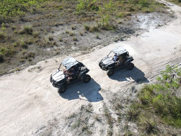 Aerial drone view of two side-by-side UTVs driving on a sandy off-road trail through grassy scrubland on a sunny day.