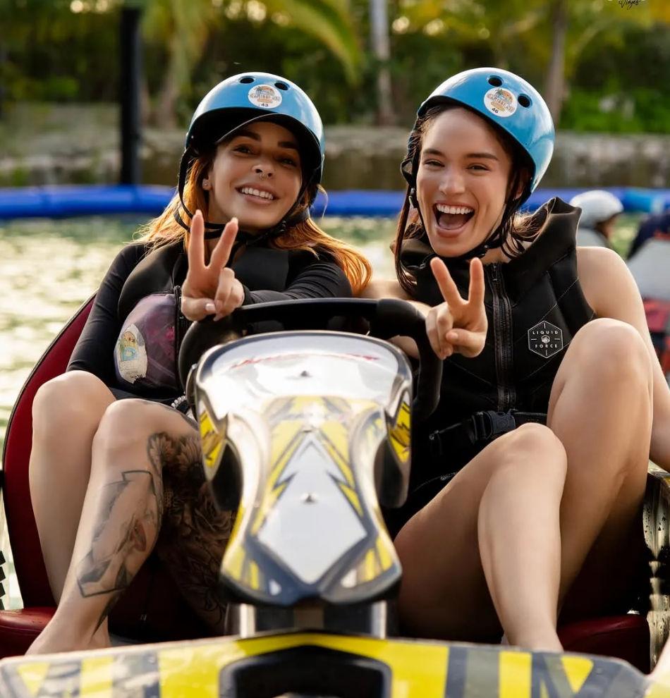 Two smiling friends in blue helmets and life vests riding a yellow jet ski on a sunny lake, flashing peace signs during a fun summer water-sports outing with green shoreline in the background.