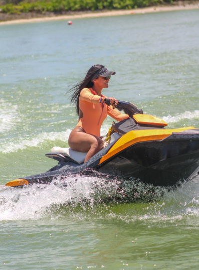Smiling woman in an orange rash guard and visor riding a yellow-and-black jet ski, splashing through green coastal waters near a tree-lined beach.