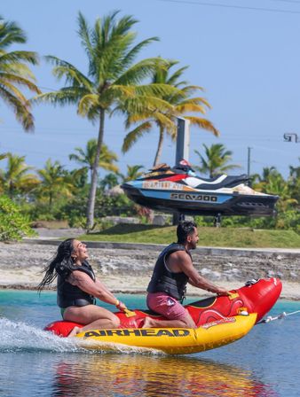 Two adults in life jackets riding a red-and-yellow inflatable towable across turquoise water near a palm-lined tropical beach, with a jet ski visible in the sunny background.
