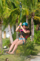 Smiling zipline rider in a teal helmet and harness gliding above a tropical palm-tree landscape
