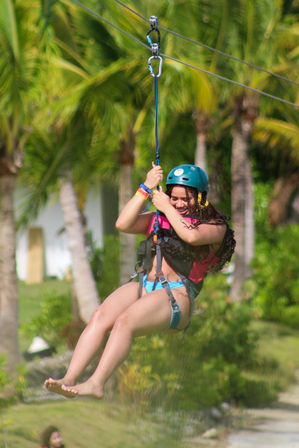 Smiling zipline rider in a teal helmet and harness gliding above a tropical palm-tree landscape