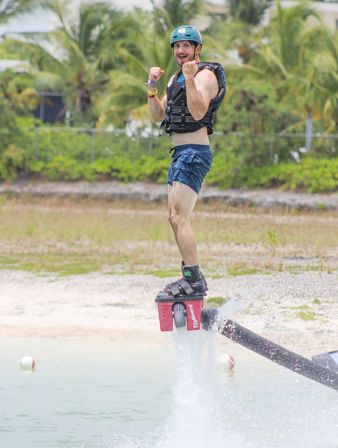 Person in helmet and life vest flyboarding above turquoise beach water, cheering with fists raised in front of palm-lined tropical shoreline