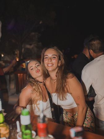 Two smiling women posing together at a lively outdoor bar at night, bottles and condiments on the wooden counter in the foreground — fun nightlife scene.