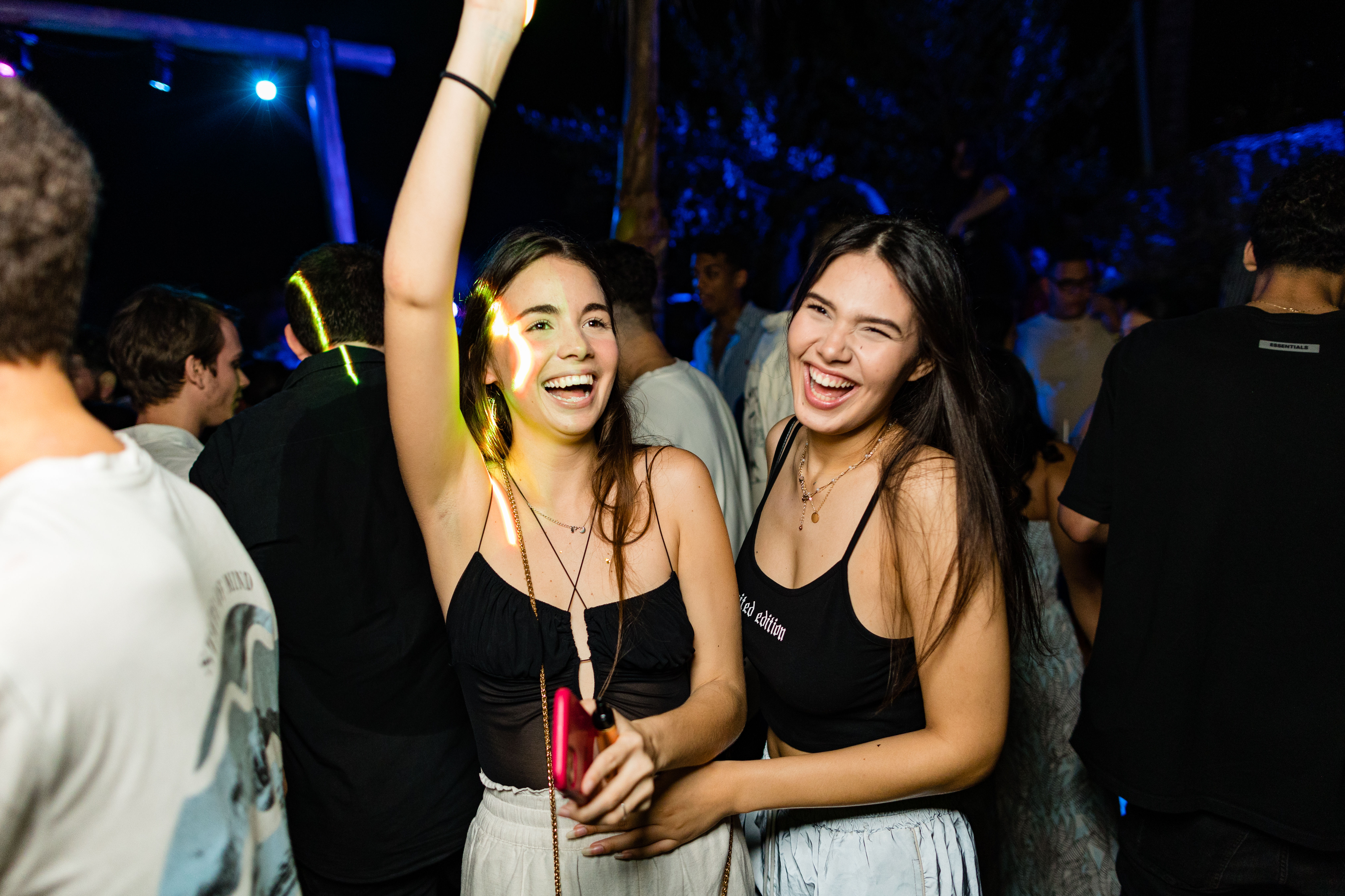 Two young women laughing and dancing together at a crowded outdoor night party under colorful lights