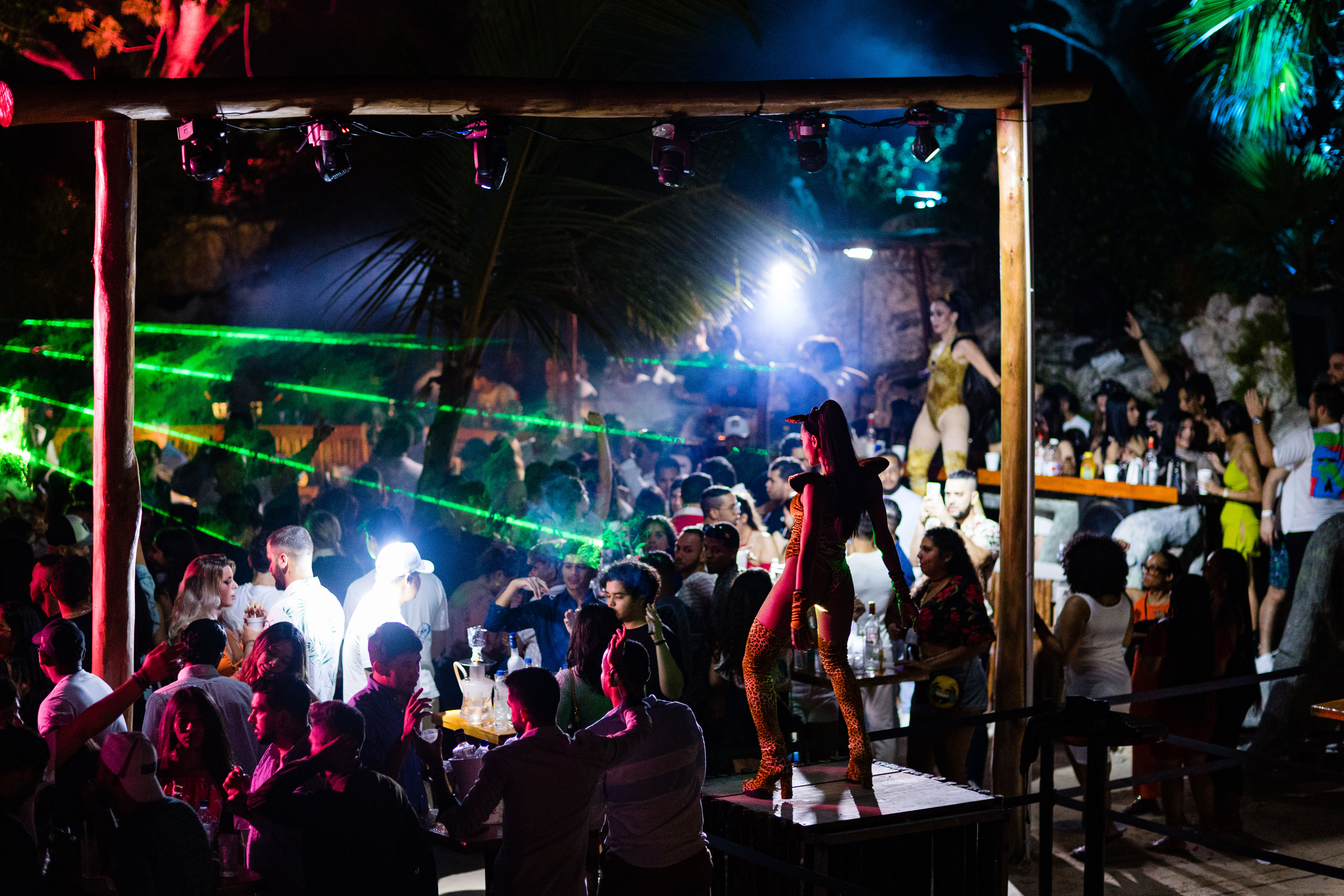 Vibrant tropical outdoor nightclub at night with green laser beams and colorful lights, a crowded dance floor and bar area, and a costumed dancer striking a pose on a raised platform.