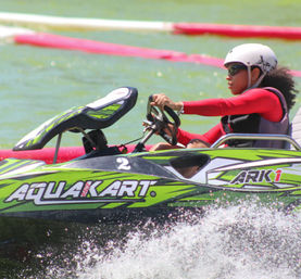 Person in white helmet and sunglasses driving a green aquatic go-kart on a lake, wearing a red long-sleeve top and life vest, gripping the steering wheel as water splashes past red course markers.