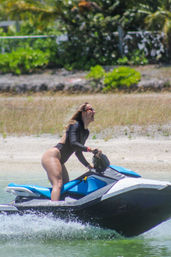 Woman in a black long-sleeve swimsuit laughing while riding a blue jet ski on clear green water near a sandy tropical shoreline with palm trees