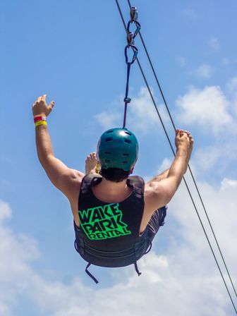 Helmeted rider in a life vest glides on a cable ride with arms raised against a bright blue summer sky at an outdoor adventure park.