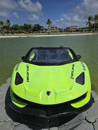 Neon lime-green sport-style jet boat docked on a floating platform in a tropical lagoon with calm turquoise water, sandy beach and palm trees under a partly cloudy blue sky.
