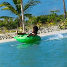 Smiling person in a black life jacket riding a bright green inflatable tube being towed across turquoise water near a tropical beach with palm trees on a sunny day
