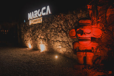 Nighttime outdoor entrance with a textured stone wall, warm ground uplights, and a red-lit carved stone totem statue framed by tropical foliage