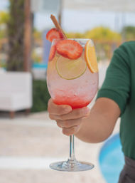 Poolside refreshing strawberry-citrus cocktail in a wine glass with lime and orange slices, strawberry garnish and cinnamon stick held by a hand.