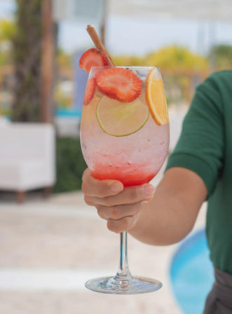 Poolside refreshing strawberry-citrus cocktail in a wine glass with lime and orange slices, strawberry garnish and cinnamon stick held by a hand.