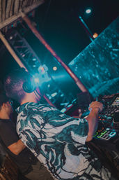 DJ wearing headphones mixes on an illuminated controller in a tropical outdoor club at night, blue stage lights and bamboo decor creating a lively nightlife vibe