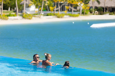 Three people relax in a sunny infinity pool overlooking a turquoise lagoon and sandy tropical beach lined with palm trees
