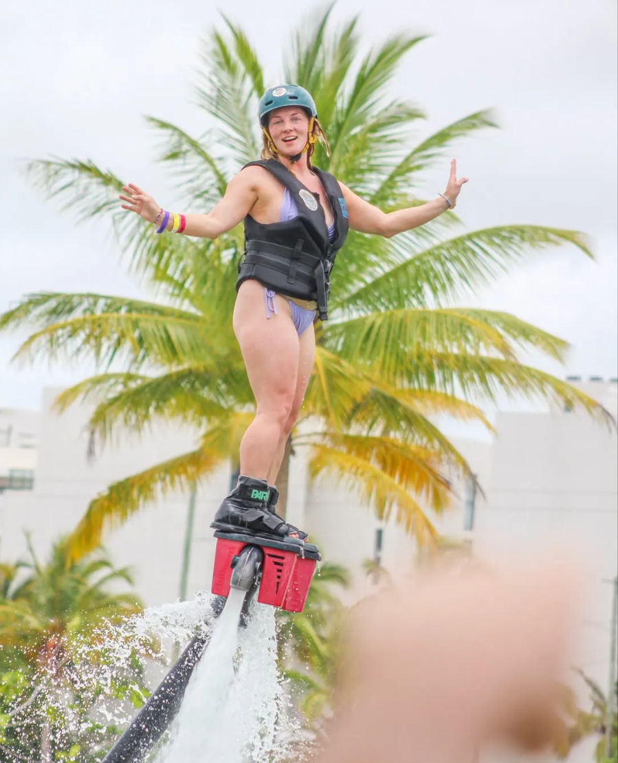 Smiling woman in a helmet and life vest riding a red flyboard water jetpack, arms outstretched above splashing water with palm trees and a tropical waterfront in the background.