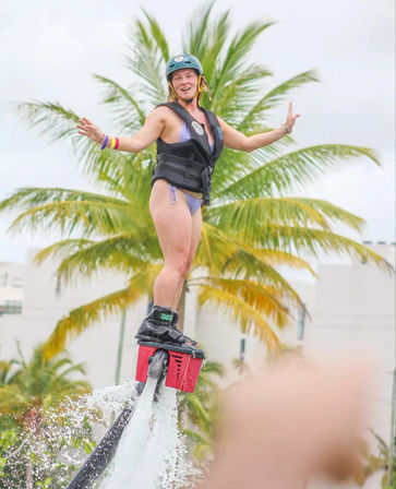 Smiling woman in a helmet and life vest riding a red flyboard water jetpack, arms outstretched above splashing water with palm trees and a tropical waterfront in the background.