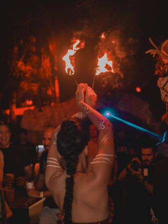 Back view of a fire performer with braided hair and white tribal body paint holding two flaming torches aloft in a dim, red-lit nightclub as a crowd and photographer watch.