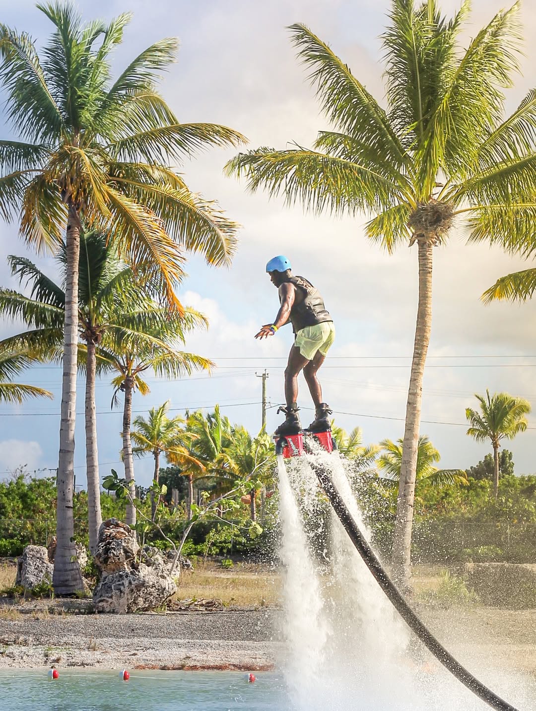 Thrilling flyboarding scene: person propelled by water jets above turquoise coastal waters with palm trees and a sunny tropical sky