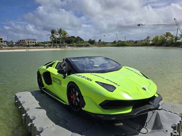 Bright lime-green convertible sports car parked on a floating dock in a shallow tropical lagoon, with palm trees, coastal buildings and cranes under a partly cloudy sky