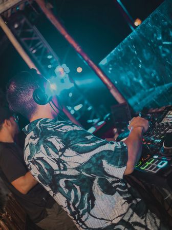 Nightclub DJ in a tropical shirt wearing headphones, hands on a mixer/controller under blue neon stage lights with crowd bokeh.