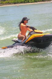 Woman in orange long-sleeve swimsuit riding a yellow and black jet ski across green coastal water, spray flying on a sunny beach day.