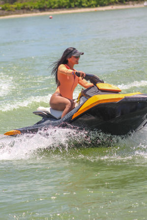Woman in orange long-sleeve swimsuit riding a yellow and black jet ski across green coastal water, spray flying on a sunny beach day.