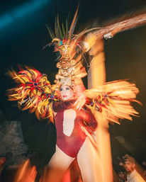 Show-stopping carnival performer in a glittering red leotard and tall feathered headdress with tiki mask, dancing under night lights with vibrant motion blur